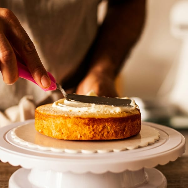 Baker spreads white frosting over a golden round cake on a decorative cake stand.