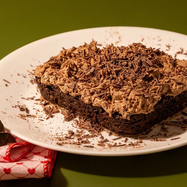 Square chocolate cake topped with chocolate frosting and shavings on a white plate beside chocolate pieces and a red heart-patterned napkin.