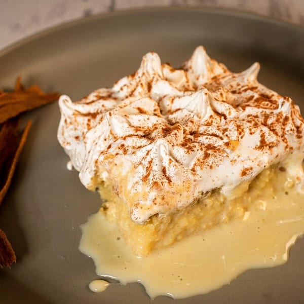 Slice of cinnamon-spiced dessert topped with whipped cream, served with cinnamon sticks on a dark plate.