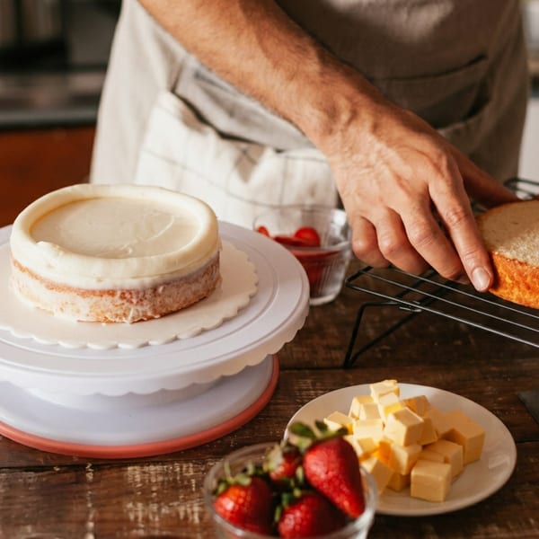 Baker assembles a layered cake beside frosting, strawberries, cheese cubes, and a cooling cake layer on a wooden table.